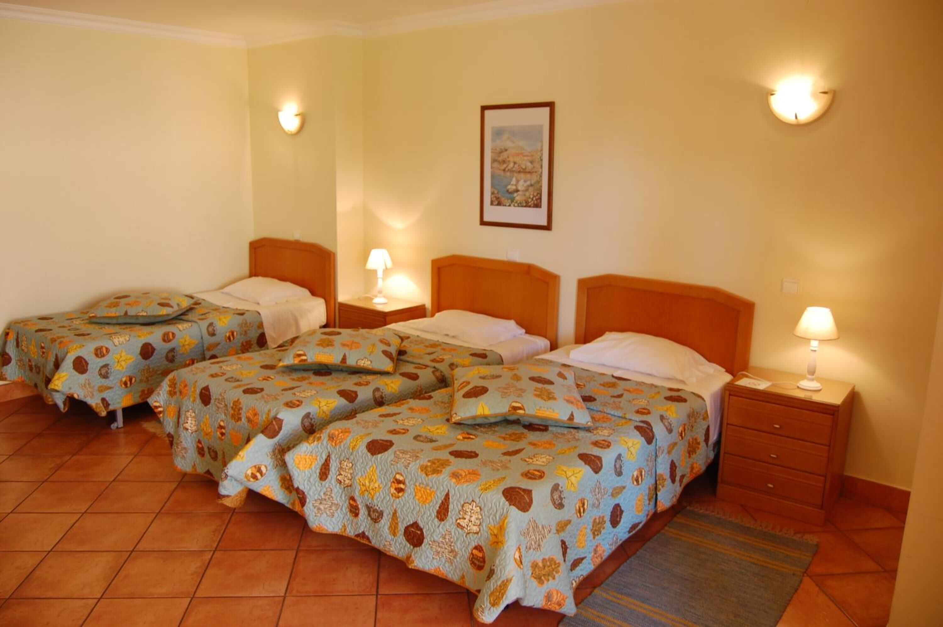 Una camera d'albergo con tre letti singoli, ognuno con copriletto e cuscini decorati. Si notano comodini con lampade, un quadro incorniciato appeso al muro e un pavimento piastrellato.