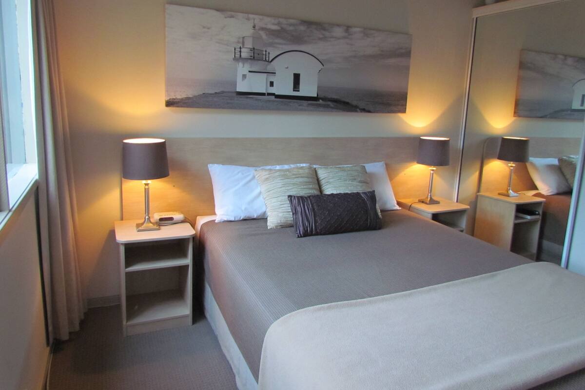 A hotel room with a bed, two bedside tables, a nightstand, a mirror, and a painting of a lighthouse on the wall.