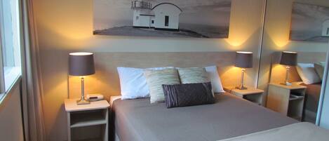 A hotel room with a bed, two bedside tables, a nightstand, a mirror, and a painting of a lighthouse on the wall.