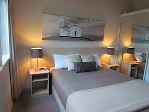 A hotel room with a bed, two bedside tables, a nightstand, a mirror, and a painting of a lighthouse on the wall.