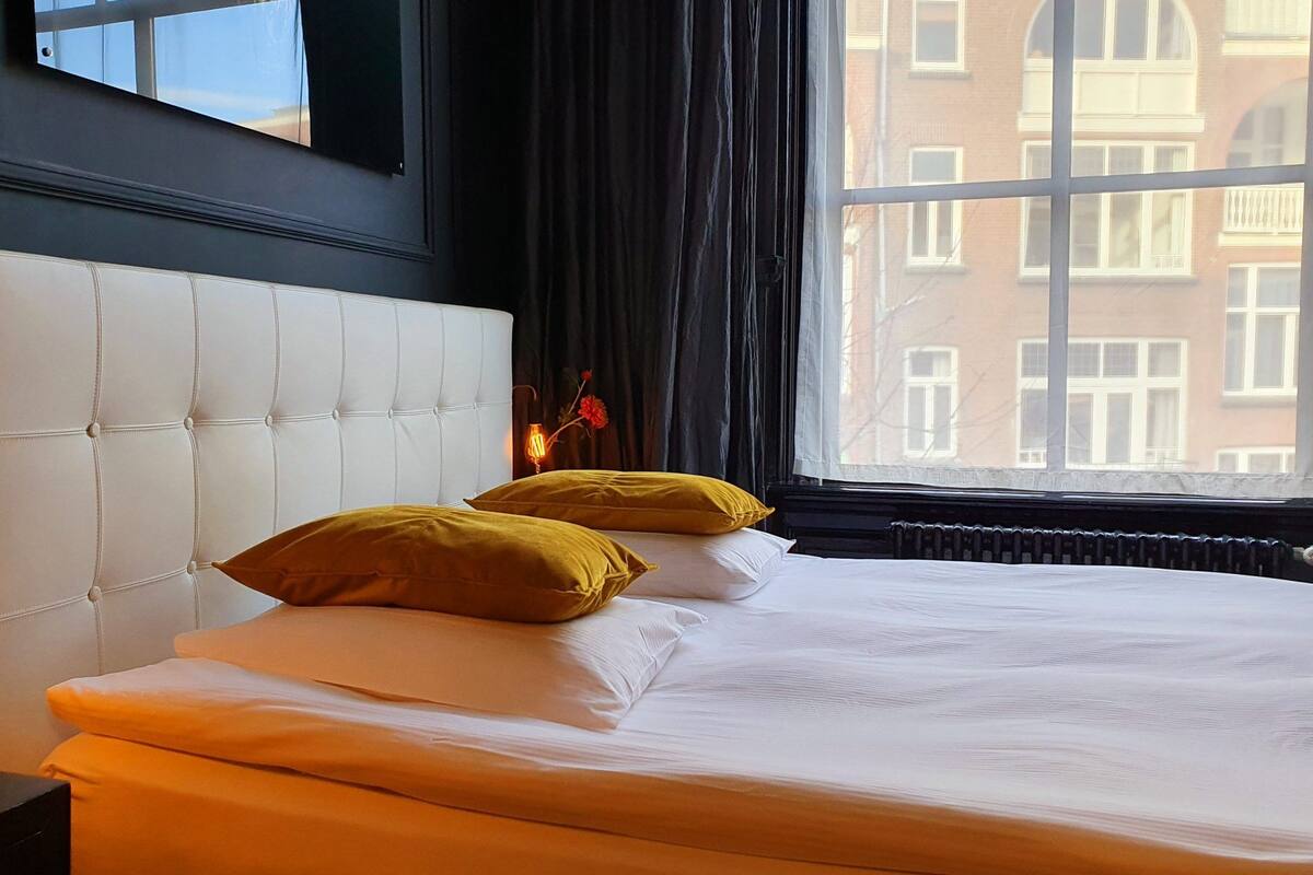 Quarto de hotel com cabeceira branca estofada, duas almofadas mostarda, colcha branca e vista de um edifício através de uma janela com cortinas escuras.