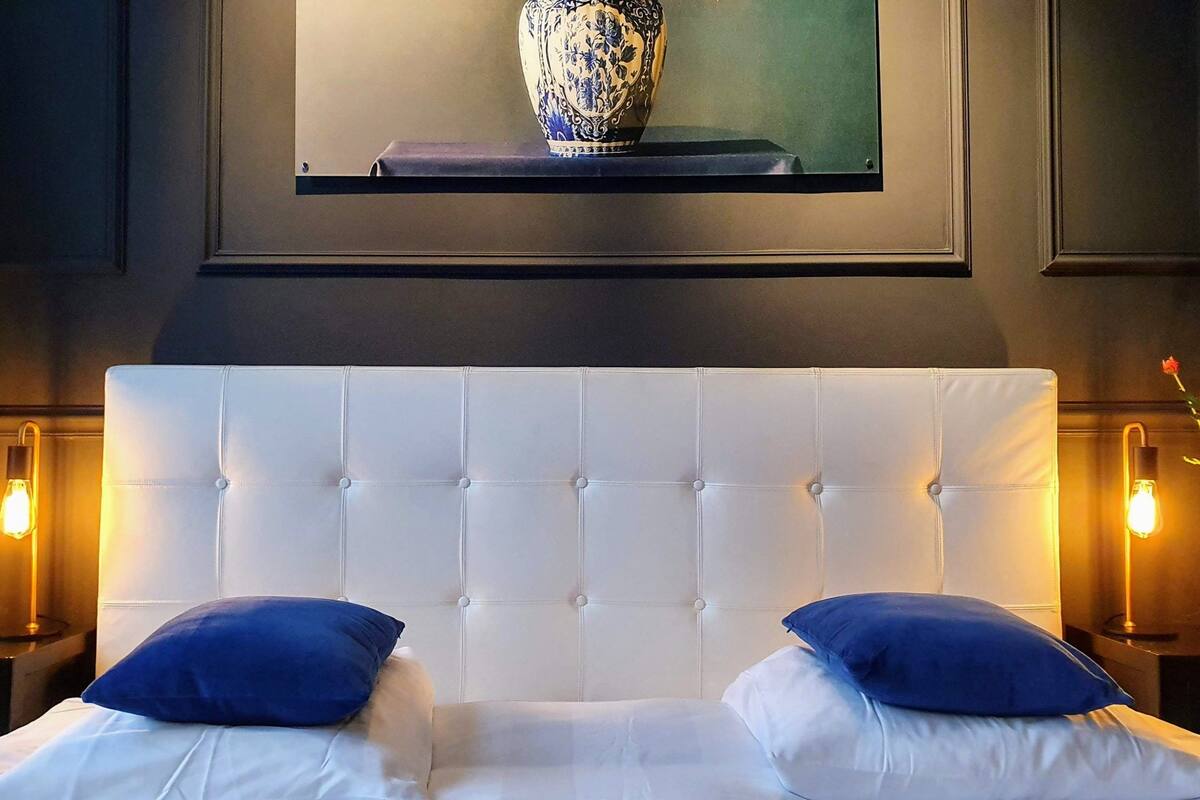 Quarto com cabeceira estofada, duas almofadas, abajures e um vaso com flores.