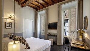 Quarto com cama, luminária de cabeceira, televisão, escrivaninha, cadeira e vista para o exterior.