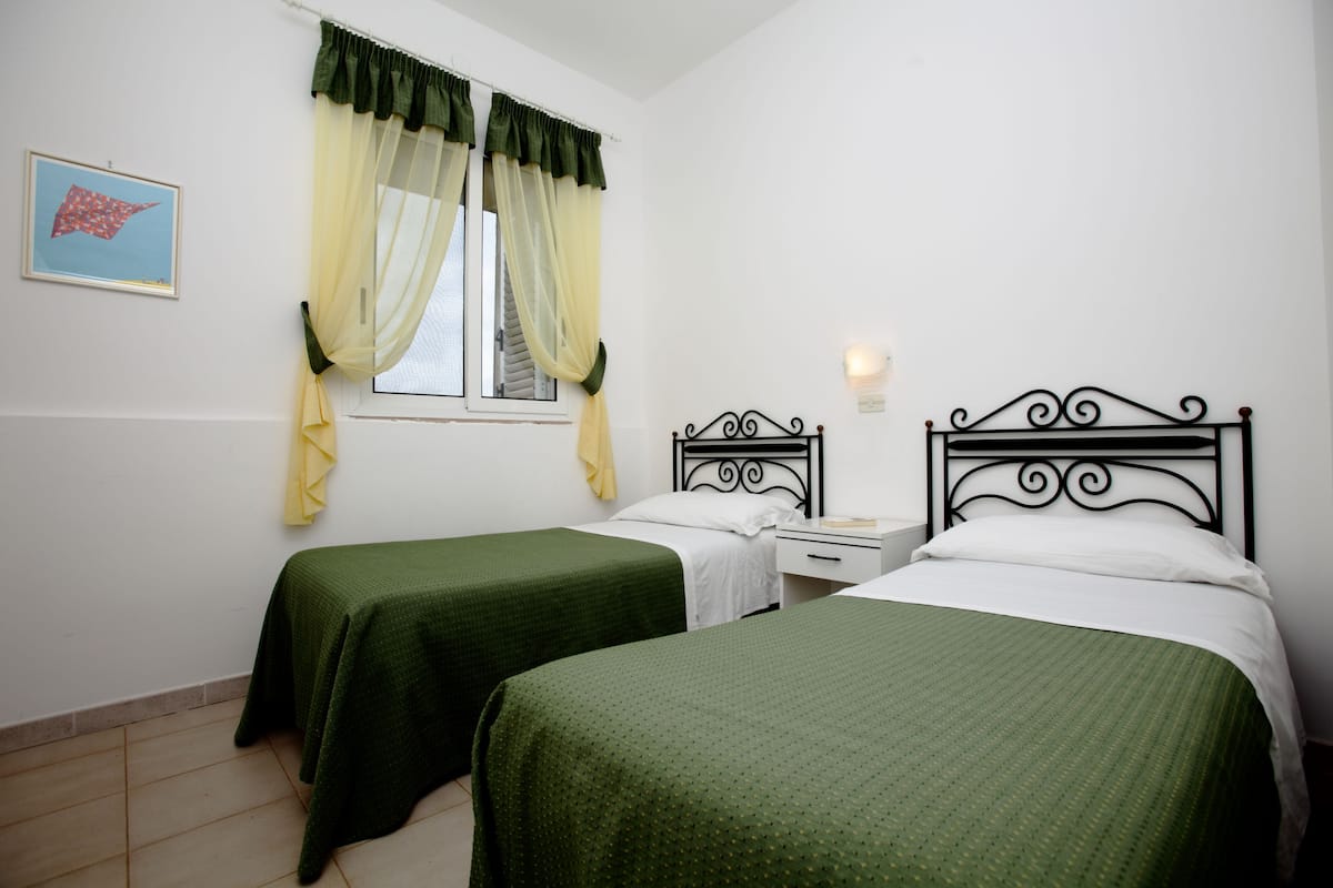 A room with two single beds, a nightstand, a window with sheer curtains, and a wall-mounted light fixture.