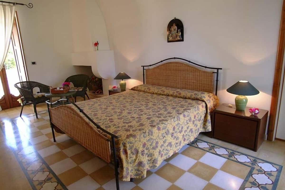 A bedroom with a checkered floor, a bed with a floral bedspread, two nightstands with lamps, a chair, and a wall-mounted decoration.