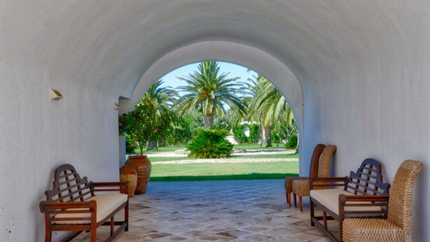 An arched corridor with a tiled floor, leading to a garden with palm trees and a lawn. There are two wooden benches and two wicker chairs along the corridor. - Masseria Tutosa (Ostuni)