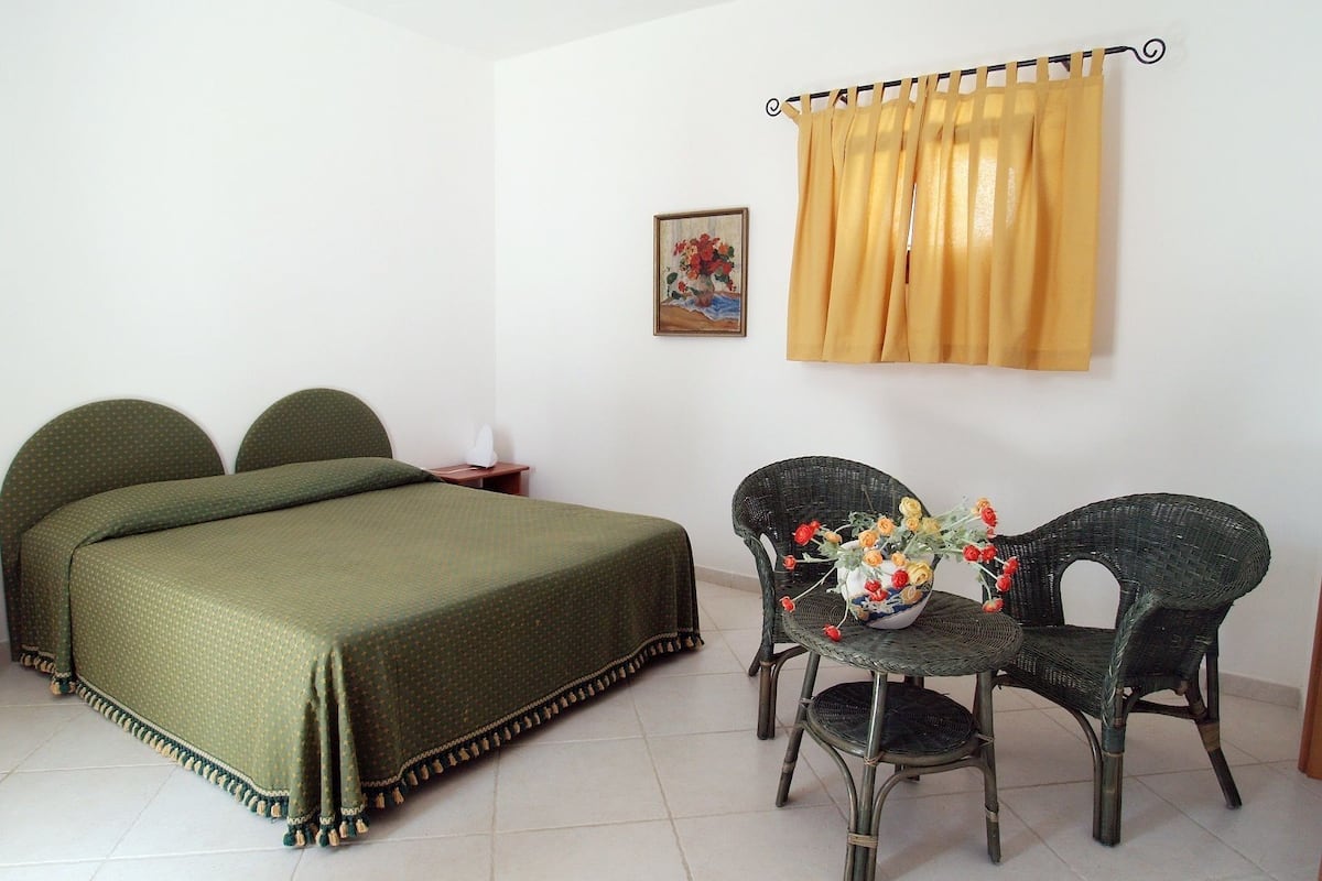 A room with a bed, two chairs, a small table with a vase of flowers, a painting on the wall, and a yellow curtain.