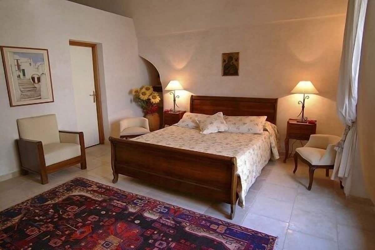 A bedroom with a wooden bed, two bedside tables with lamps, a chair, a rug, and a painting on the wall.