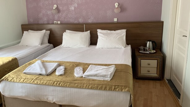 Habitación de hotel con dos camas individuales, una cama doble y una mesita de noche de madera con tetera y un cuadro enmarcado.