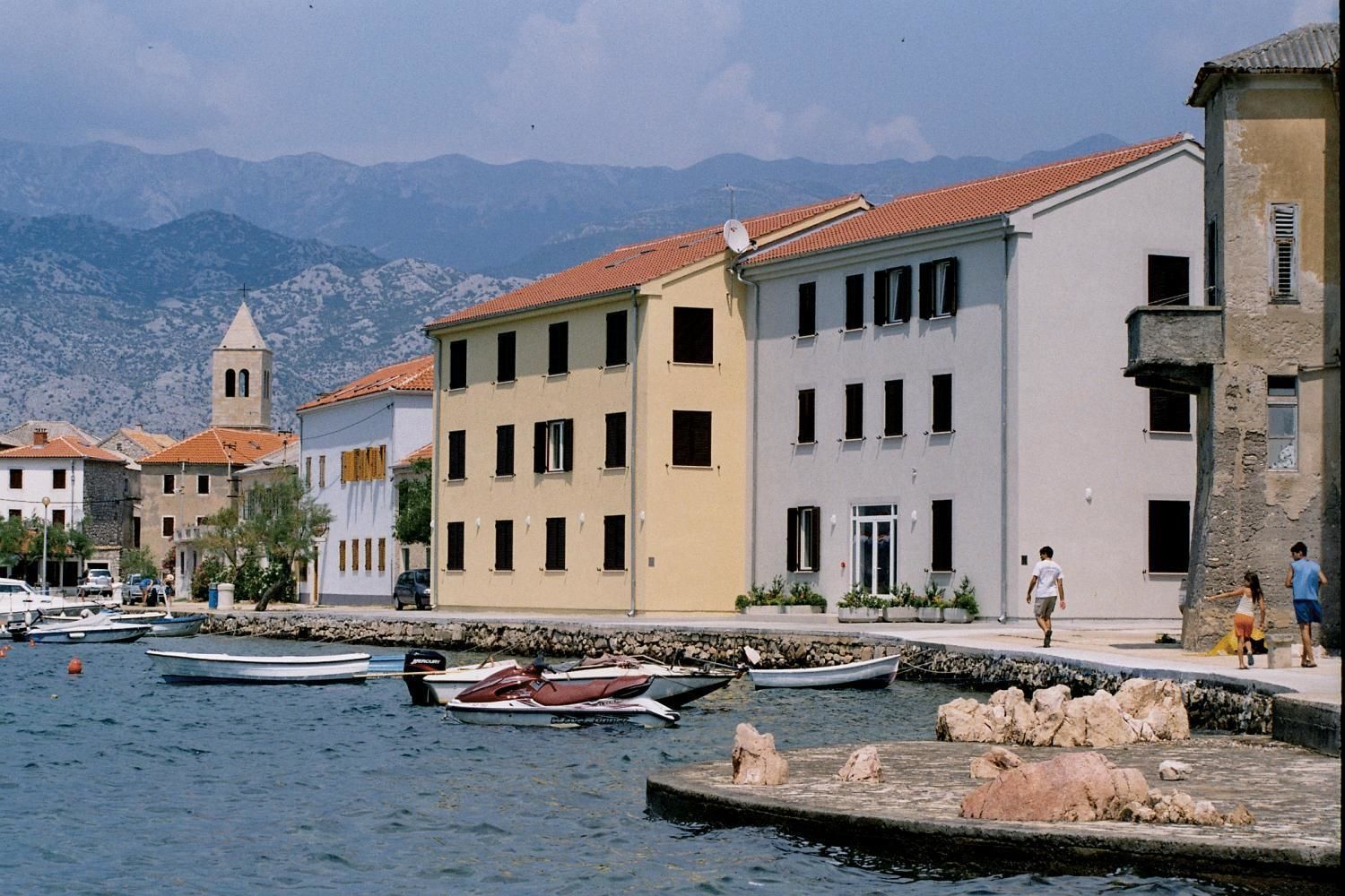 Un pueblo costero con edificios a lo largo del malecón, una iglesia con campanario y barcos amarrados en el muelle.