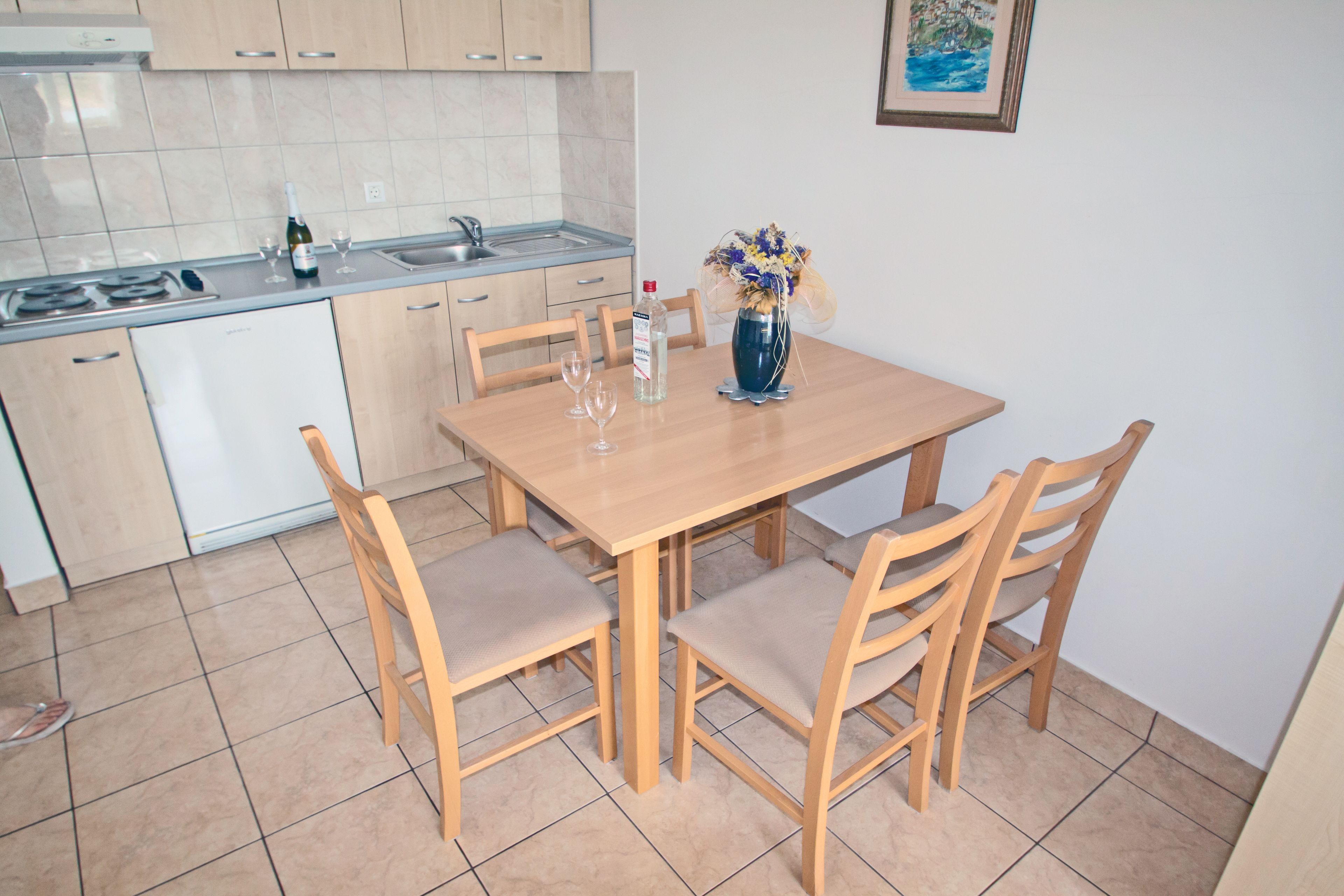 Una cocina con una mesa de comedor, sillas, un florero con flores, una botella de agua y una copa de vino.