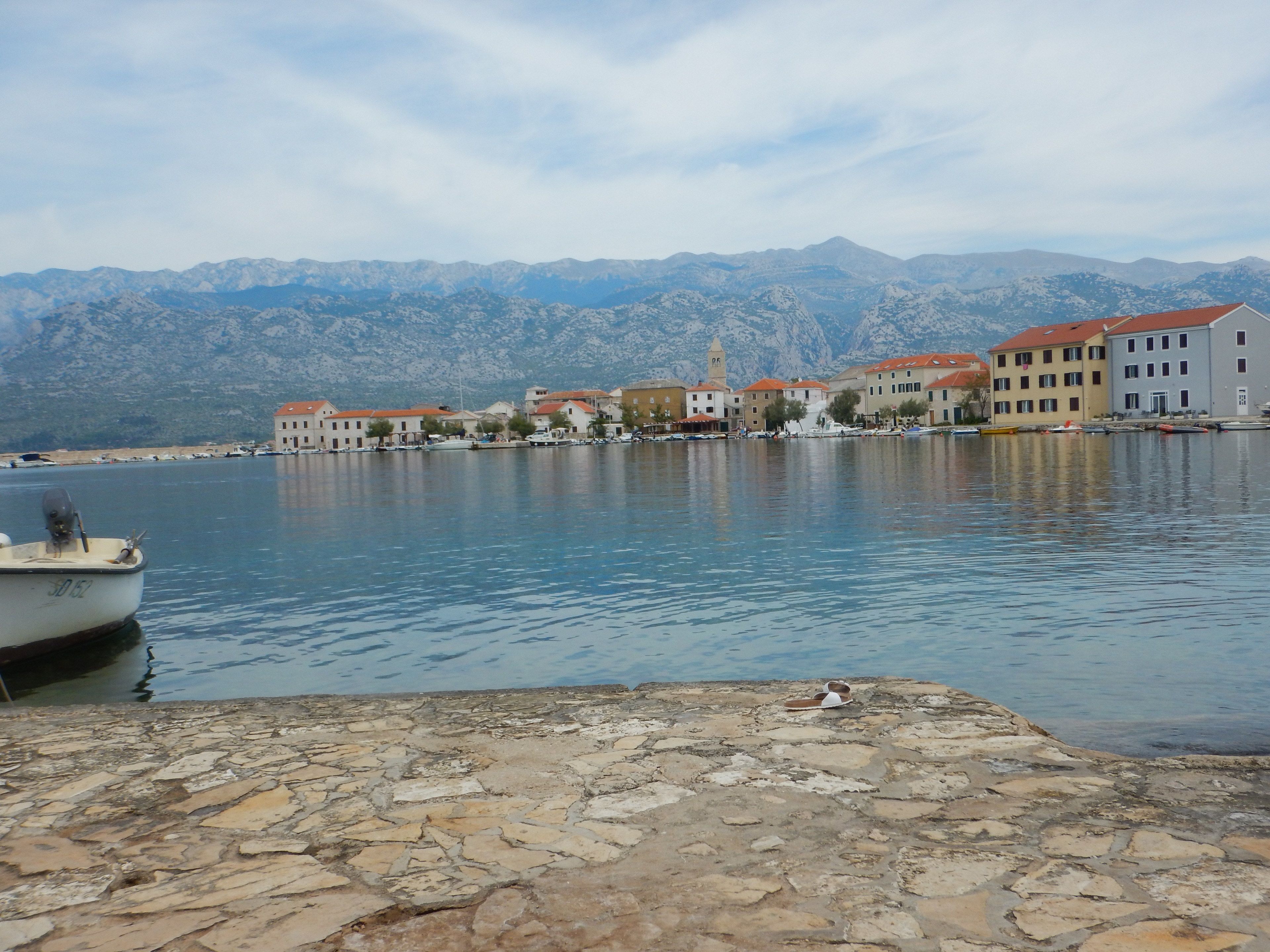Vista sul lungomare con banchina in pietra, acqua calma e edifici con tetti rossi sullo sfondo.