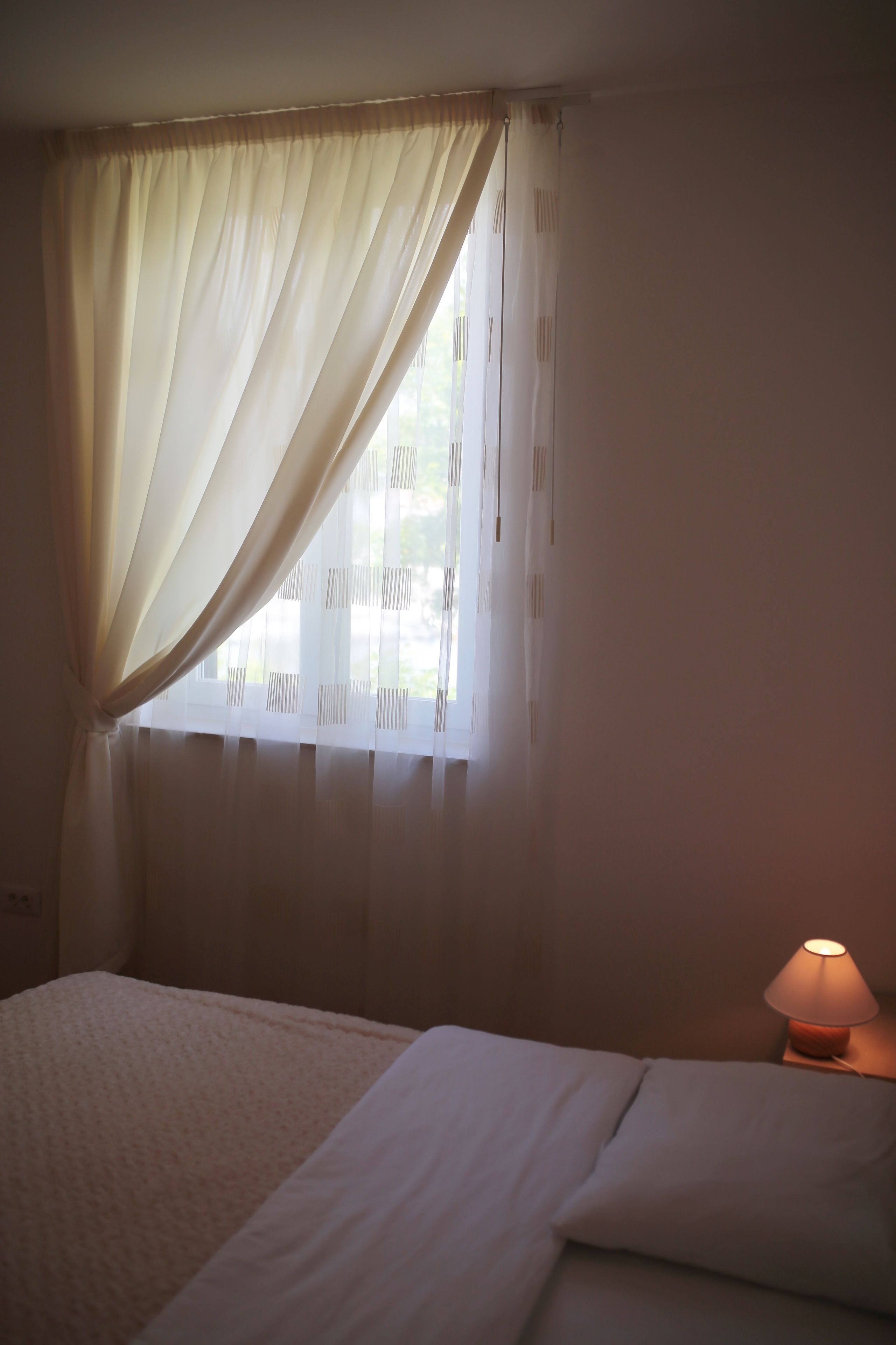 A bedroom with a bed, a window with sheer curtains, and a bedside lamp.