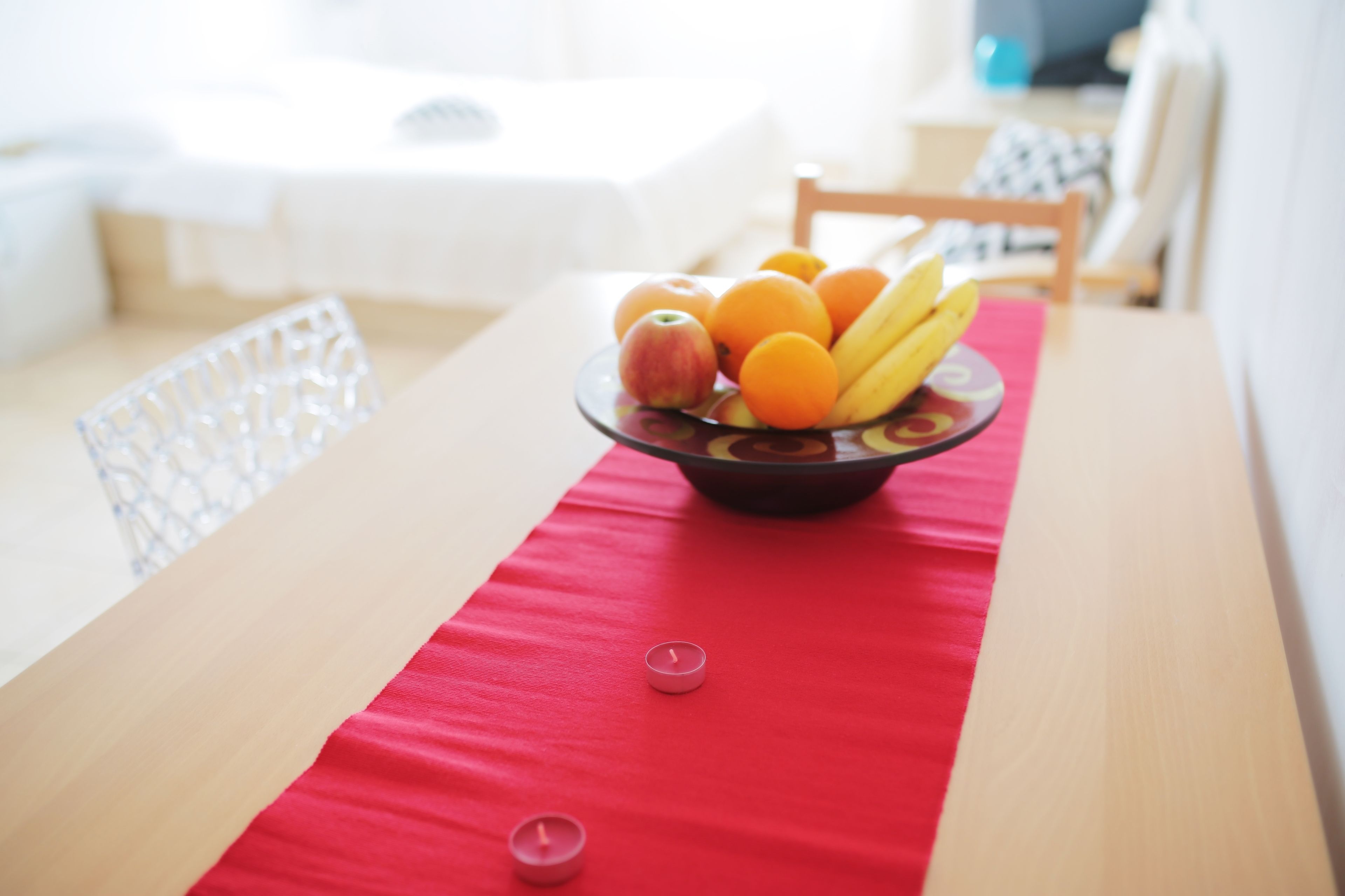 Une table à manger ornée d’un chemin de table rouge, d’un saladier rempli de fruits et de deux bougies allumées.