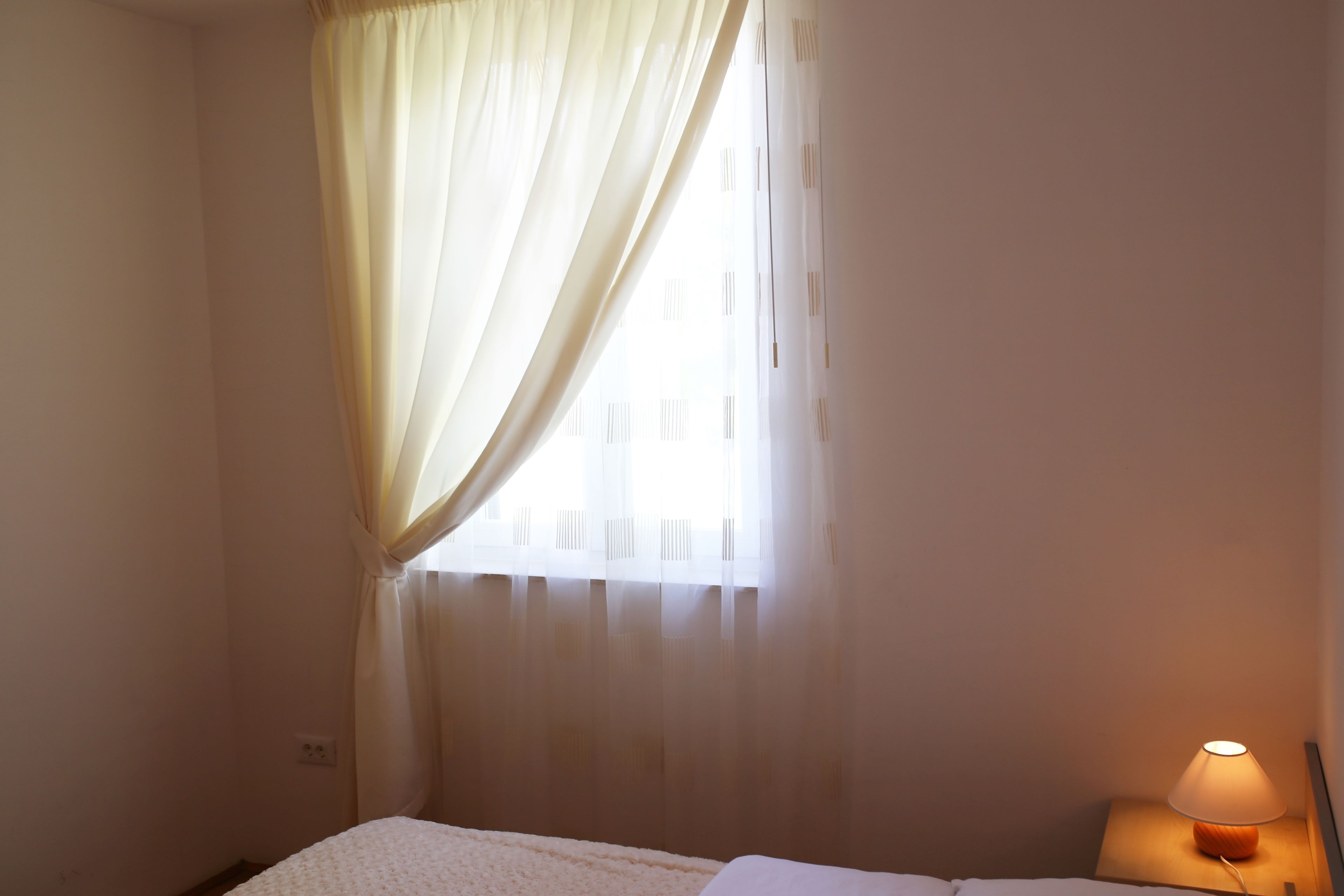 A hotel room with a bed, a nightstand with a lamp, sheer curtains, and a window with blinds.