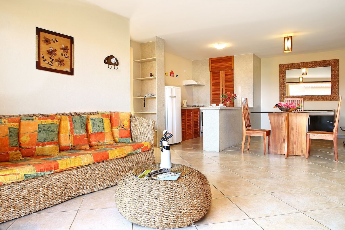 A living room with a sofa, ottoman, and a kitchen area in the background.