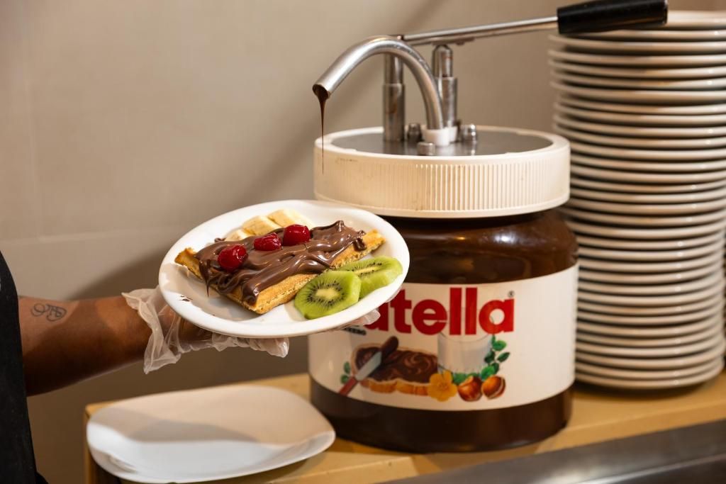 A person holding a plate with a dessert topped with chocolate and fruit, a jar of Nutella, and a stack of plates.