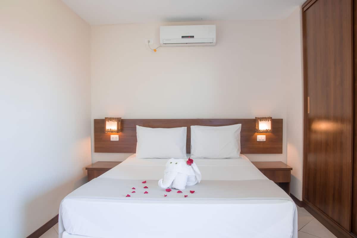 A hotel room with a bed, two bedside lamps, a wooden wardrobe, and an air conditioning unit.