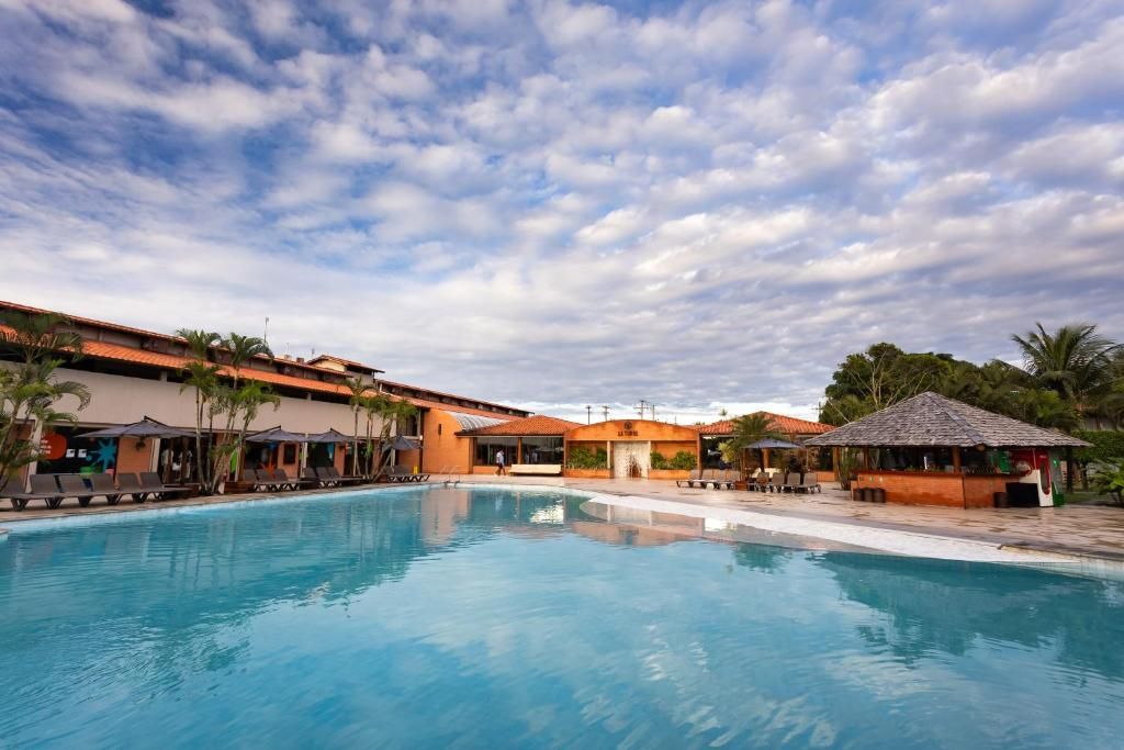 Piscina com espreguiçadeiras e guarda-sóis, cercada por edifícios com arquitetura tropical.