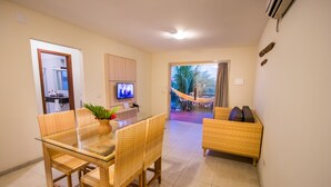 A modern living room with a glass dining table, wicker furniture, a TV, and a view of a hammock and palm trees outside. - La Torre Resort All Inclusive (Porto Seguro)