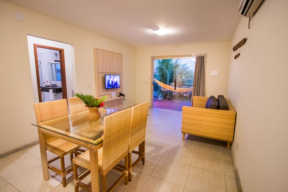 A modern living room with a glass dining table, wicker furniture, a TV, and a view of a hammock and palm trees outside.