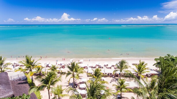 A beach with clear turquoise water, palm trees, and beach umbrellas. - La Torre Resort All Inclusive (Porto Seguro)