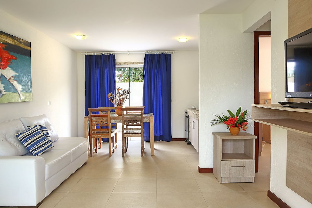 A modern living room with a white sofa, a dining table with chairs, a TV, and a kitchen area with a cabinet and a small plant.