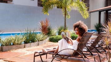 Una persona sentada en una silla de descanso leyendo un libro en un área exterior bien cuidada, con una piscina y diversas plantas.