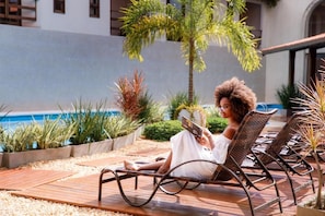A person sitting on a lounge chair reading a book in a well-maintained outdoor area with a pool and various plants. - La Torre Resort All Inclusive (Porto Seguro)