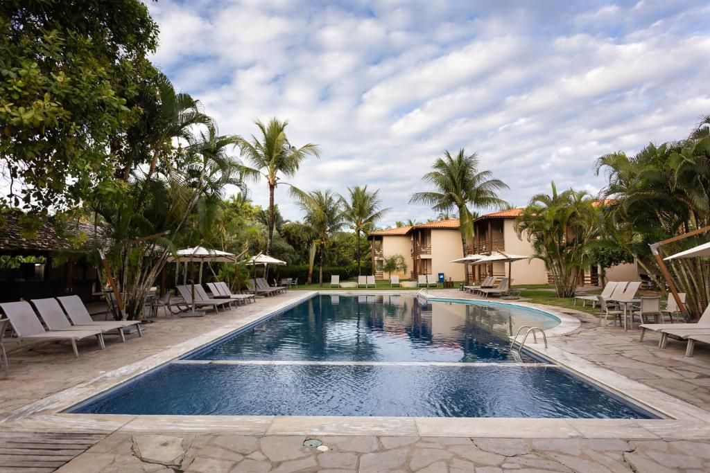 A swimming pool with lounge chairs and umbrellas, surrounded by palm trees and a building with a tiled roof.