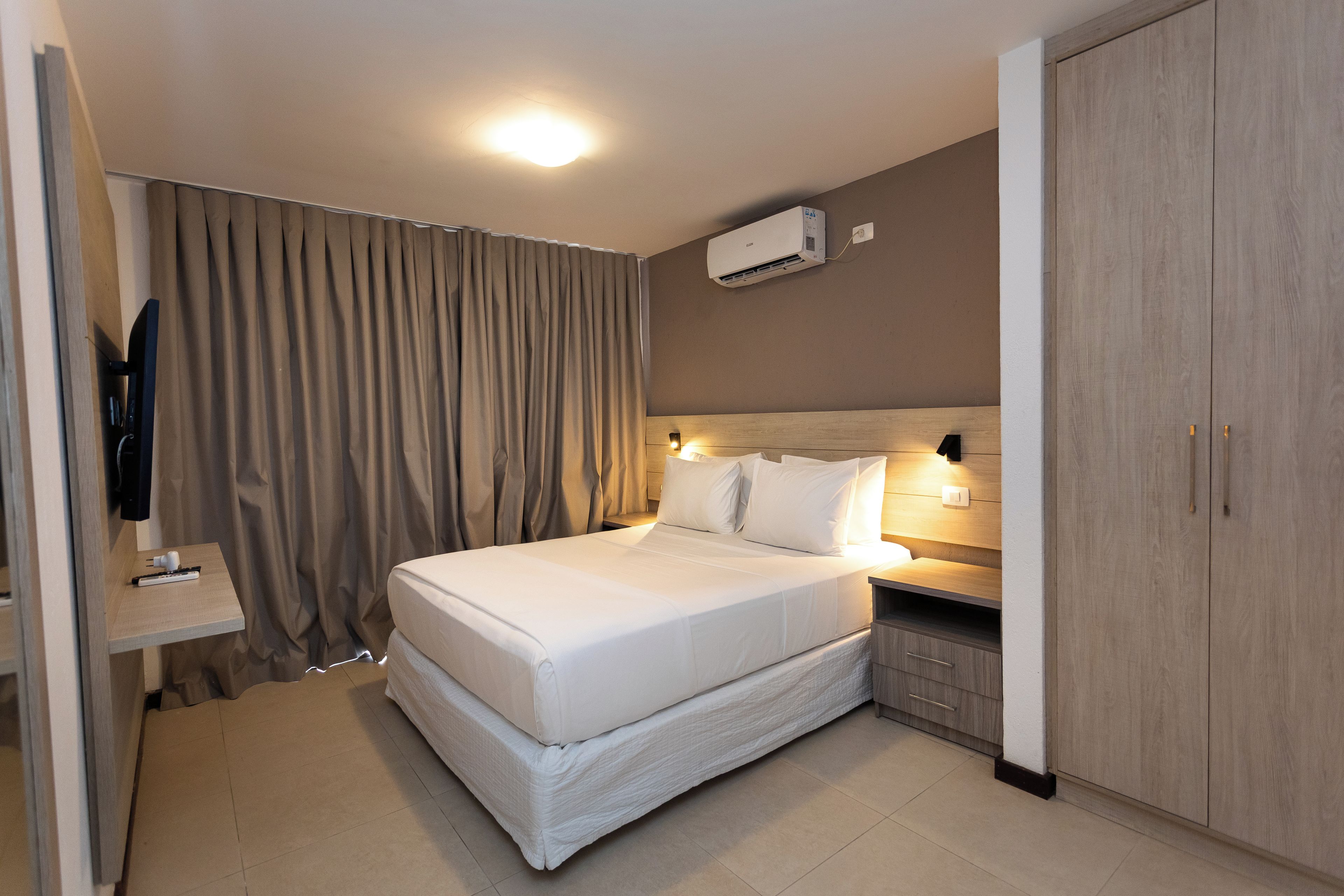 A hotel room with a bed, a bedside table, a wardrobe, and an air conditioning unit.