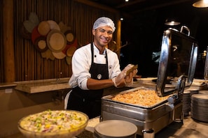 Un bufet ben a punt, amb un cuiner servint menjar, una gran safata de metall i una pizza en un bol.
