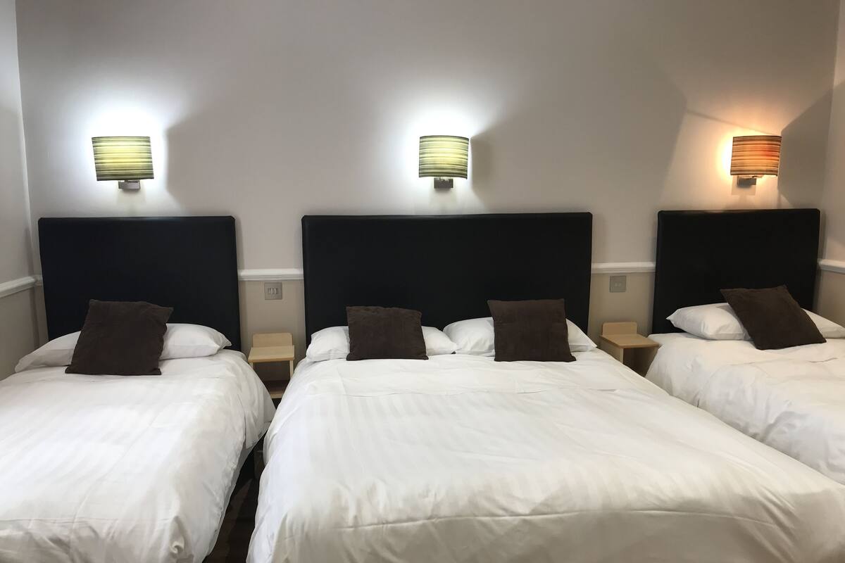 Trois lits simples avec des têtes de lit sombres et une literie blanche, chacun étant équipé d’une table de chevet et d’une lampe fixée au mur.