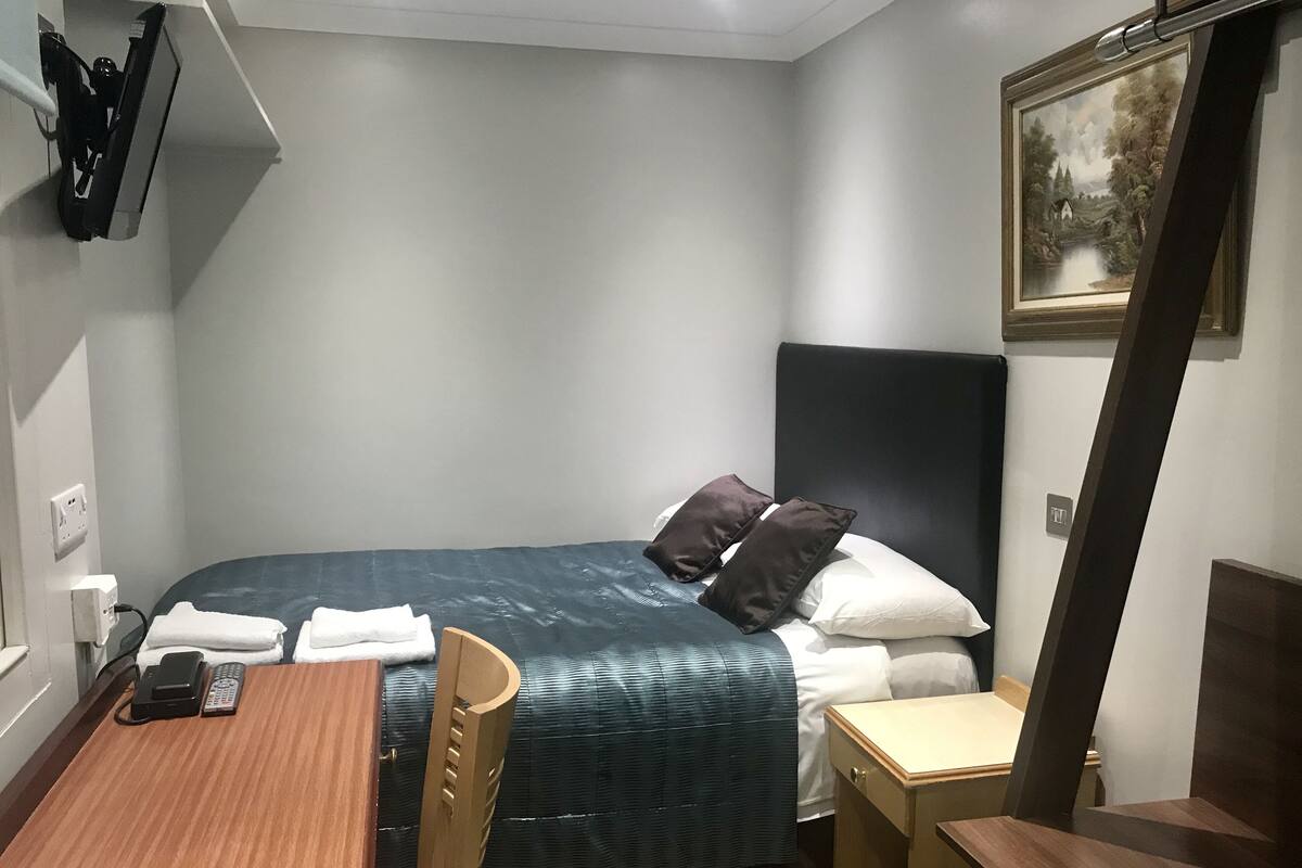 Une chambre d’hôtel de taille modeste, comprenant un lit simple, un bureau en bois, une chaise et une petite table.