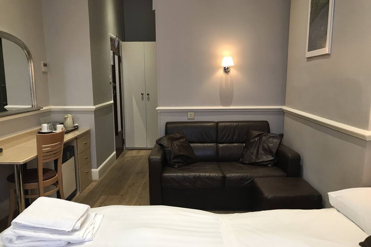 Une chambre d’hôtel comprenant un lit, un bureau, une chaise, un canapé en cuir, une armoire et un luminaire fixé au mur.