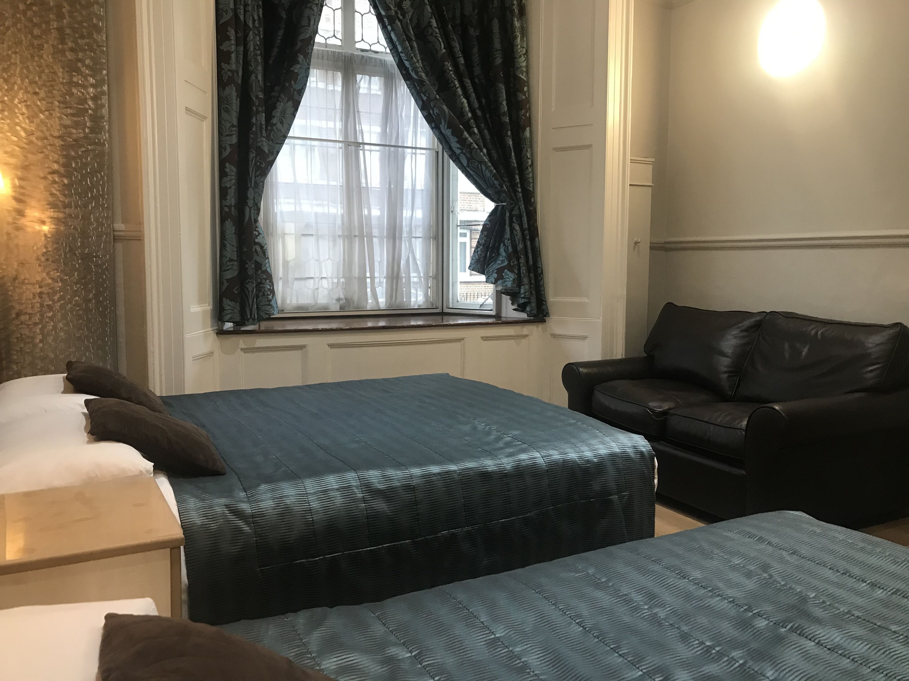 A room with two beds, a black leather sofa, a window with curtains, and a wall-mounted light.