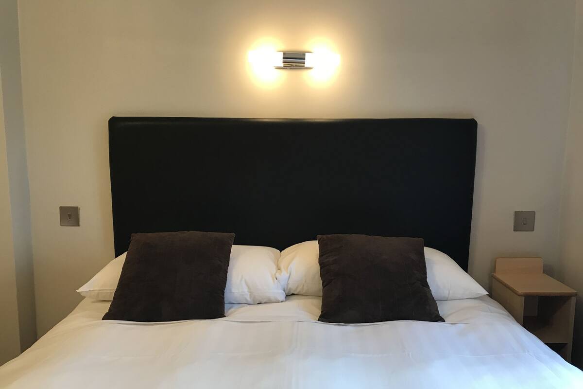 Un lit bien fait, avec du linge de lit blanc et deux oreillers foncés, une tête de lit noire et un luminaire fixé au mur.