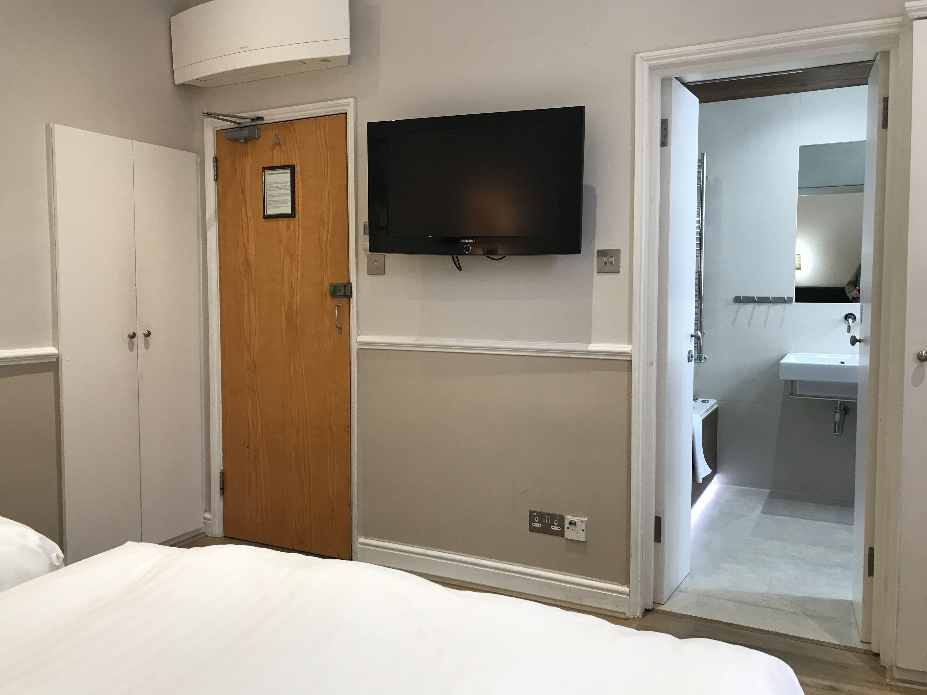 Una habitación de hotel con cama, puerta de madera, televisor montado y baño con lavamanos y espejo.