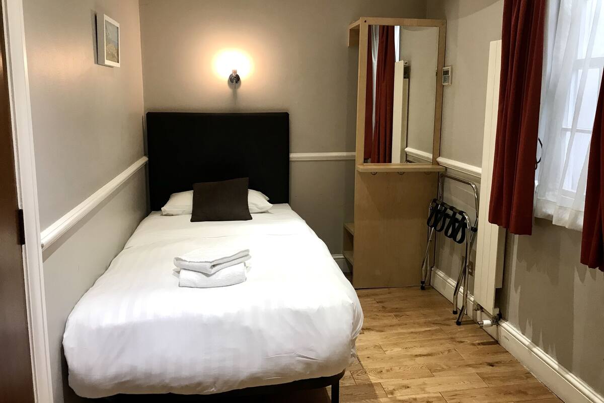 Un lit bien fait, avec du linge de lit blanc et un seul oreiller marron, un luminaire fixé au mur, une armoire en bois avec un miroir et un radiateur posé sur le sol.