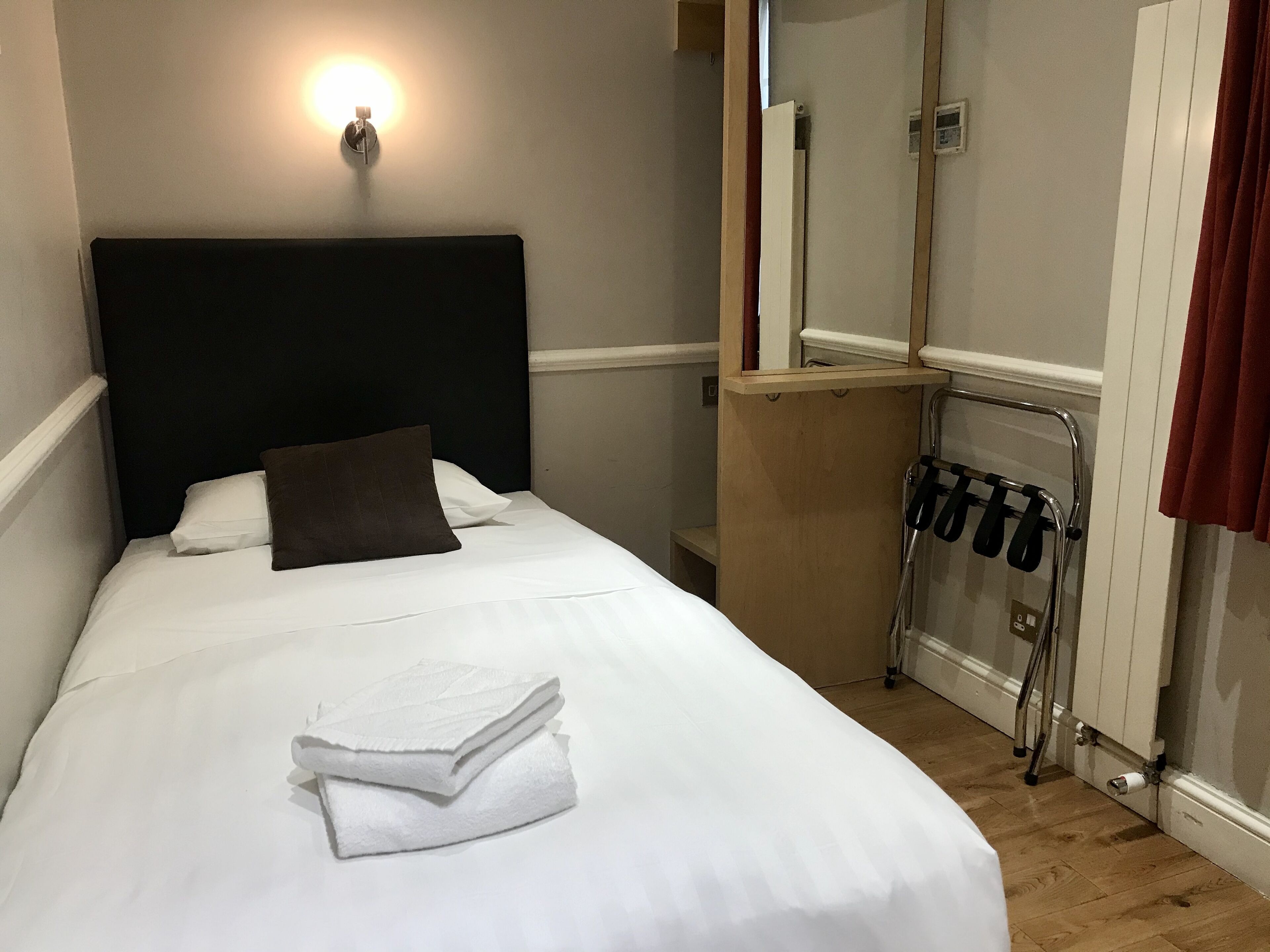 A neatly made bed with white linens and a brown pillow, a wooden headboard, a wall-mounted light, and a mirror above the bed.