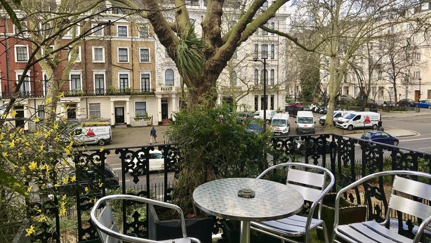 A terrace with metal chairs and a table, overlooking a street with parked cars and buildings.