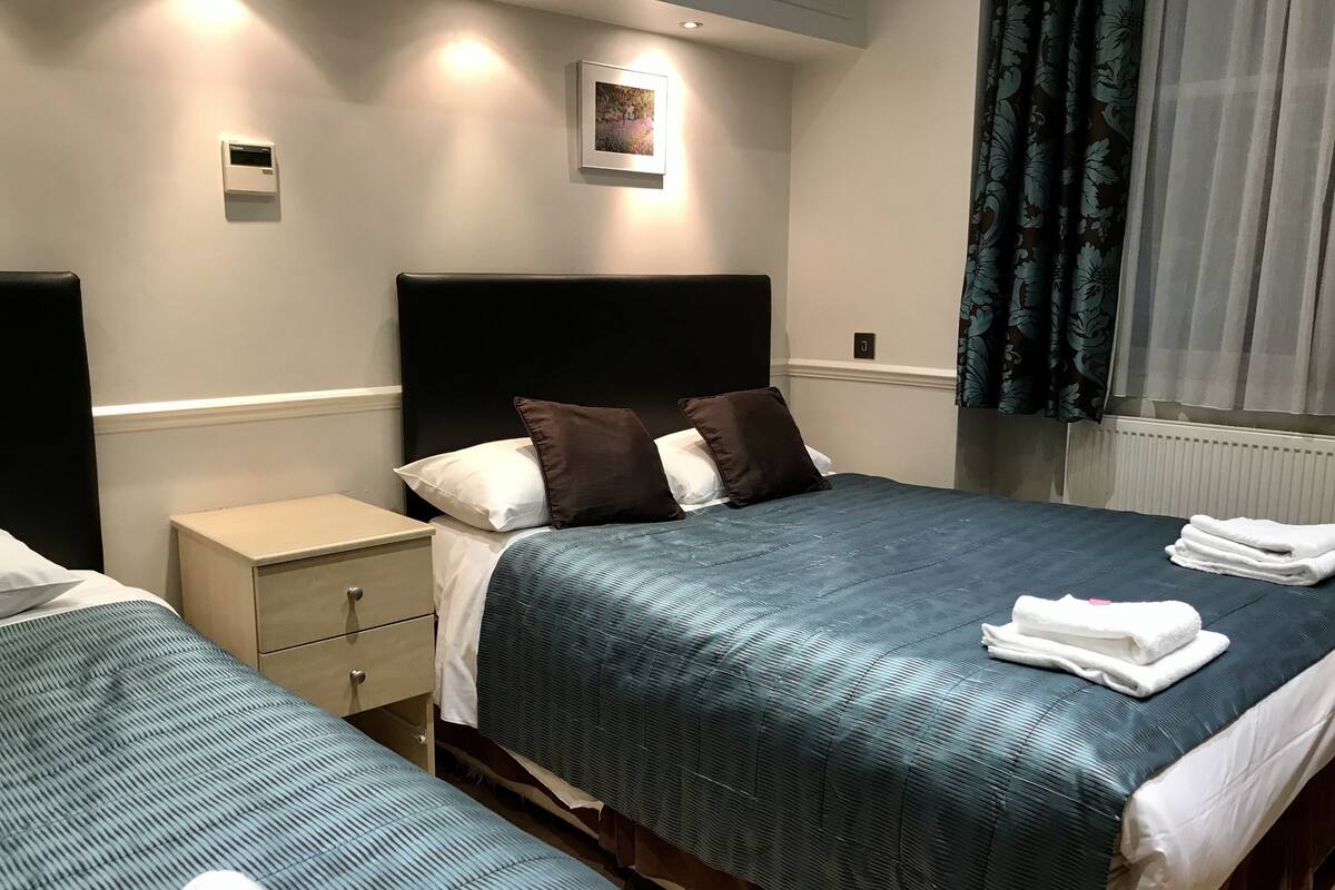 Une chambre d’hôtel avec deux lits, chacun recouvert d’une couverture bleue et agrémenté d’oreillers blancs. On y trouve une table de chevet avec un tiroir, un radiateur fixé au mur et un tableau encadré au-dessus de la tête de lit.