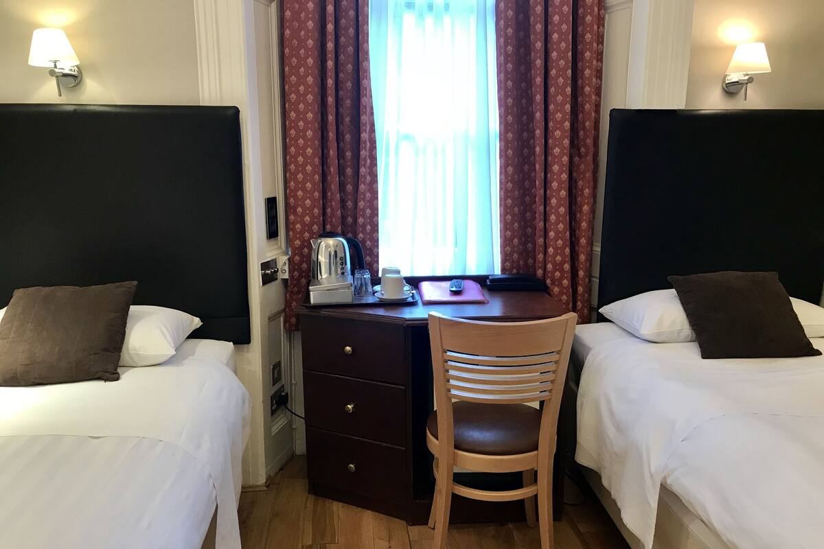 Une chambre d’hôtel avec deux lits simples, un bureau avec une chaise, une bouilloire et une fenêtre avec des rideaux.