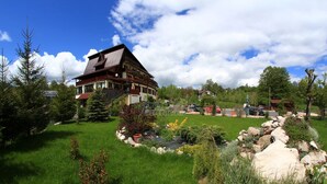 Uma casa grande e de vários andares com telhado inclinado, rodeada por um jardim bem cuidado com várias plantas e um muro de pedra.