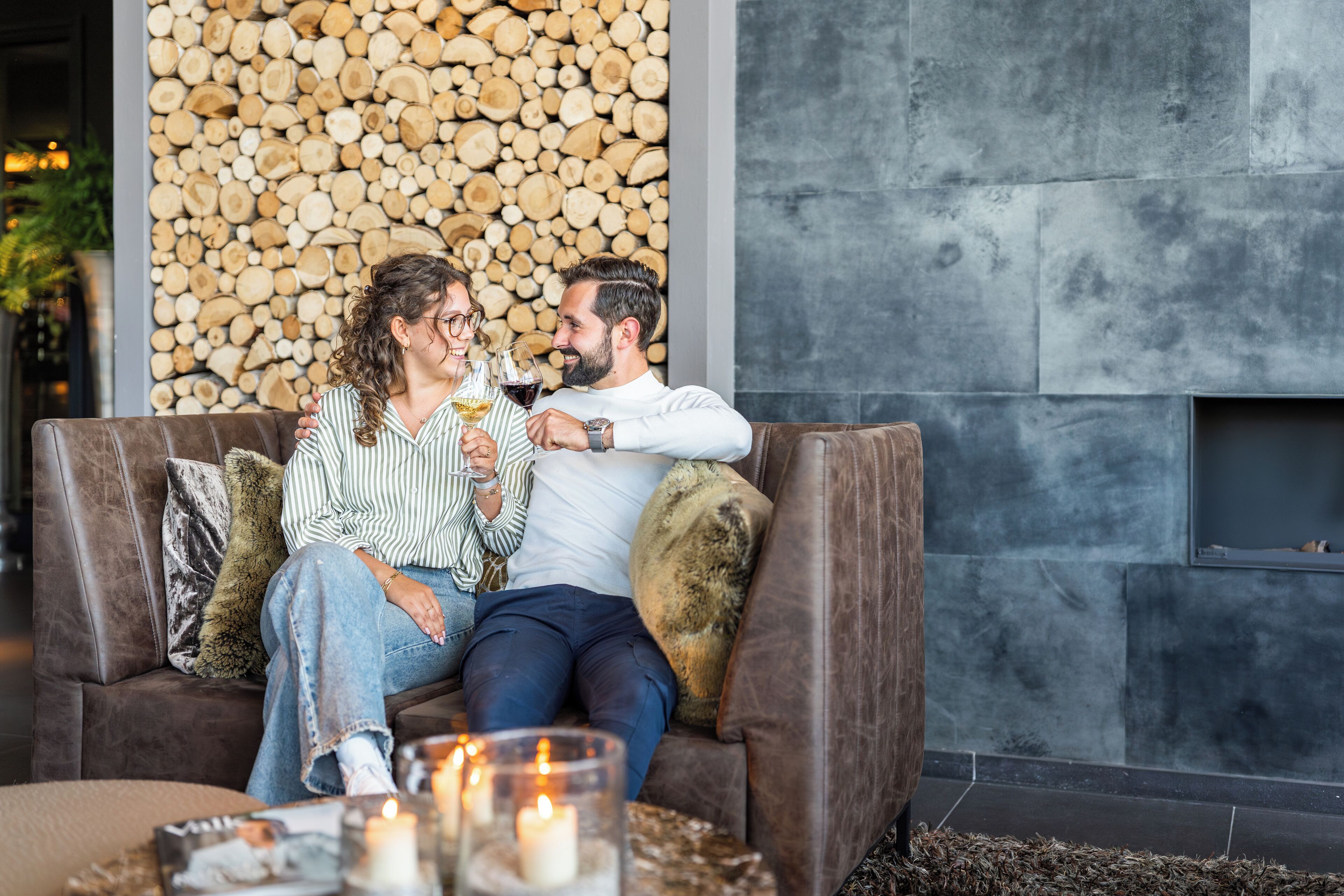 A couple sitting on a brown leather sofa with a stack of logs in the background, a fireplace, and candles on a side table.