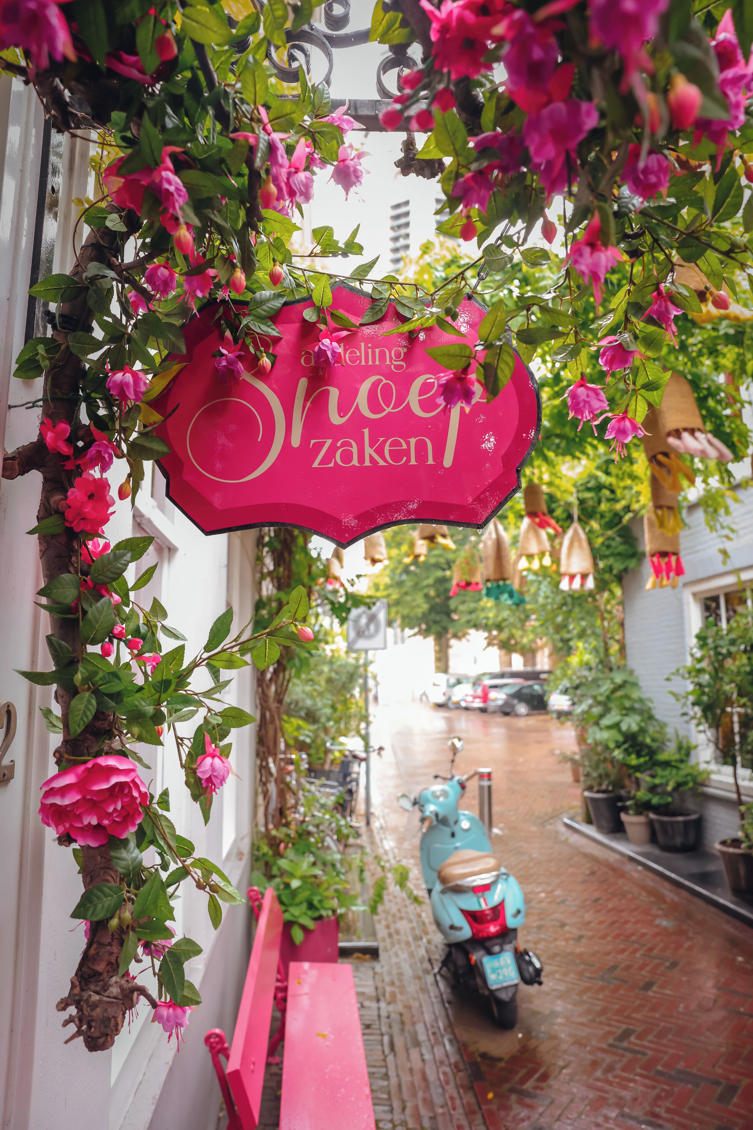 A narrow street with a scooter parked on the side, a bench, and a pink sign hanging from a tree.