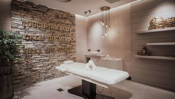 Uma sala de spa moderna com parede de pedra, mesa de massagem, pia e prateleiras com objetos decorativos.