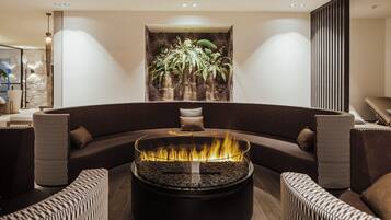 Un espace de détente moderne avec un foyer circulaire, des canapés aux couleurs sombres et une grande plante fixée au mur.