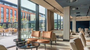 Un espace de détente intérieur moderne, doté de murs en verre, offrant une vue sur un bâtiment rouge et aménagé avec un mélange de fauteuils en cuir et en tissu.