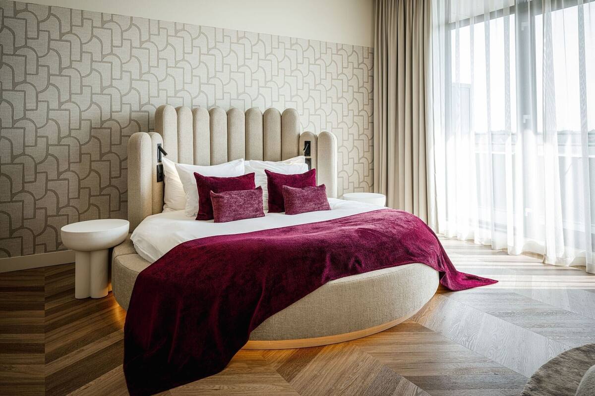 Een moderne slaapkamer met een wand met een geometrisch patroon, een groot bed met hoofdeinde en uitzicht op het buitengebeuren door dunne gordijnen.